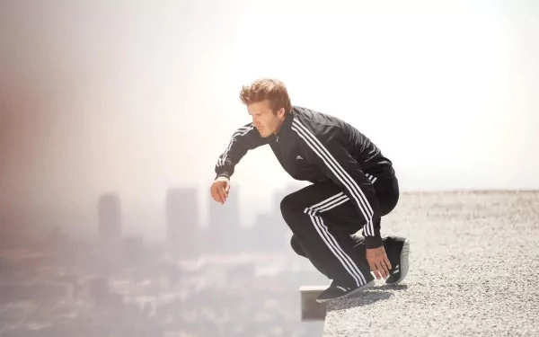 A striking HD desktop wallpaper featuring David Beckham in a sleek black tracksuit, poised in a dynamic crouch against a city skyline backdrop, embodying the spirit of sports excellence.
