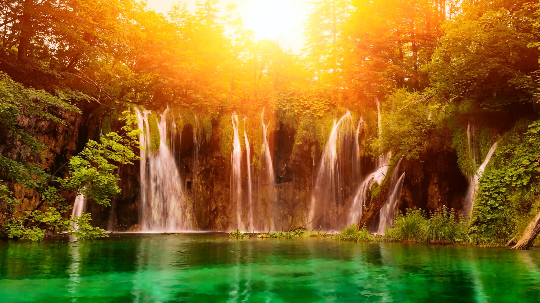 A stunning HD desktop wallpaper showcasing a vibrant waterfall cascading into a serene green pool, surrounded by lush trees and bathed in warm sunlight.