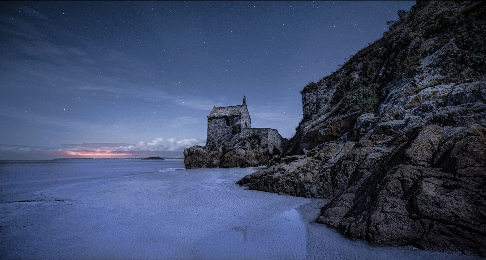 A serene HD desktop wallpaper featuring a stone ruin by the sea, illuminated by twilight, surrounded by rocky terrain and a calm, reflective shoreline.
