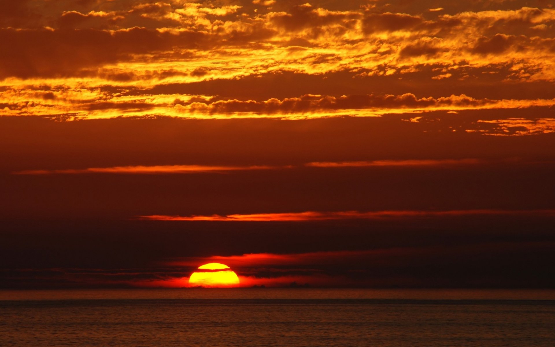 HD PC desktop wallpaper featuring a vibrant nature sunset with the sun partially visible above the horizon and glowing orange clouds in the sky.
