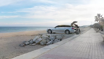 HD desktop wallpaper featuring a silver Jaguar XF parked near a rocky beach with a person unloading a bicycle beside a paved walkway.