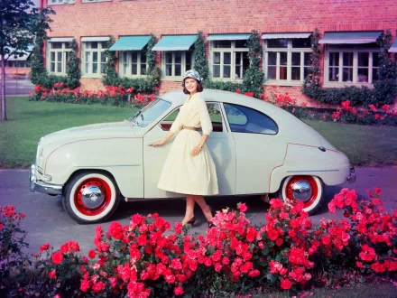 Woman in a pale dress beside a cream 1959 Saab 93B classic vehicle parked in a flowered front yard — 2K Quad HD PC desktop wallpaper background.