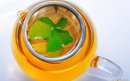 A close-up of a glass teapot filled with golden tea, featuring fresh green leaves steeping inside, creating a serene and refreshing food and tea-themed wallpaper.