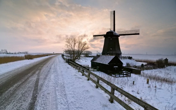 A snow-covered landscape features a traditional windmill beside a winding road, set against a dramatic sky, creating a stunning HD desktop wallpaper and background.