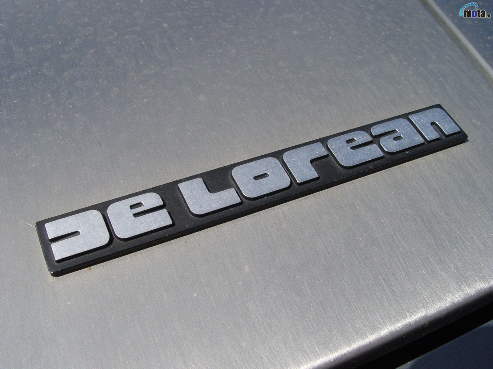 Close-up of DeLorean vehicle emblem on brushed stainless steel — HD PC desktop wallpaper background.