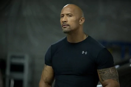 Dwayne Johnson stands confidently in a fitted black shirt, showcasing his muscular build. The background features a dimly lit gym setting, enhancing the focus on his powerful presence.