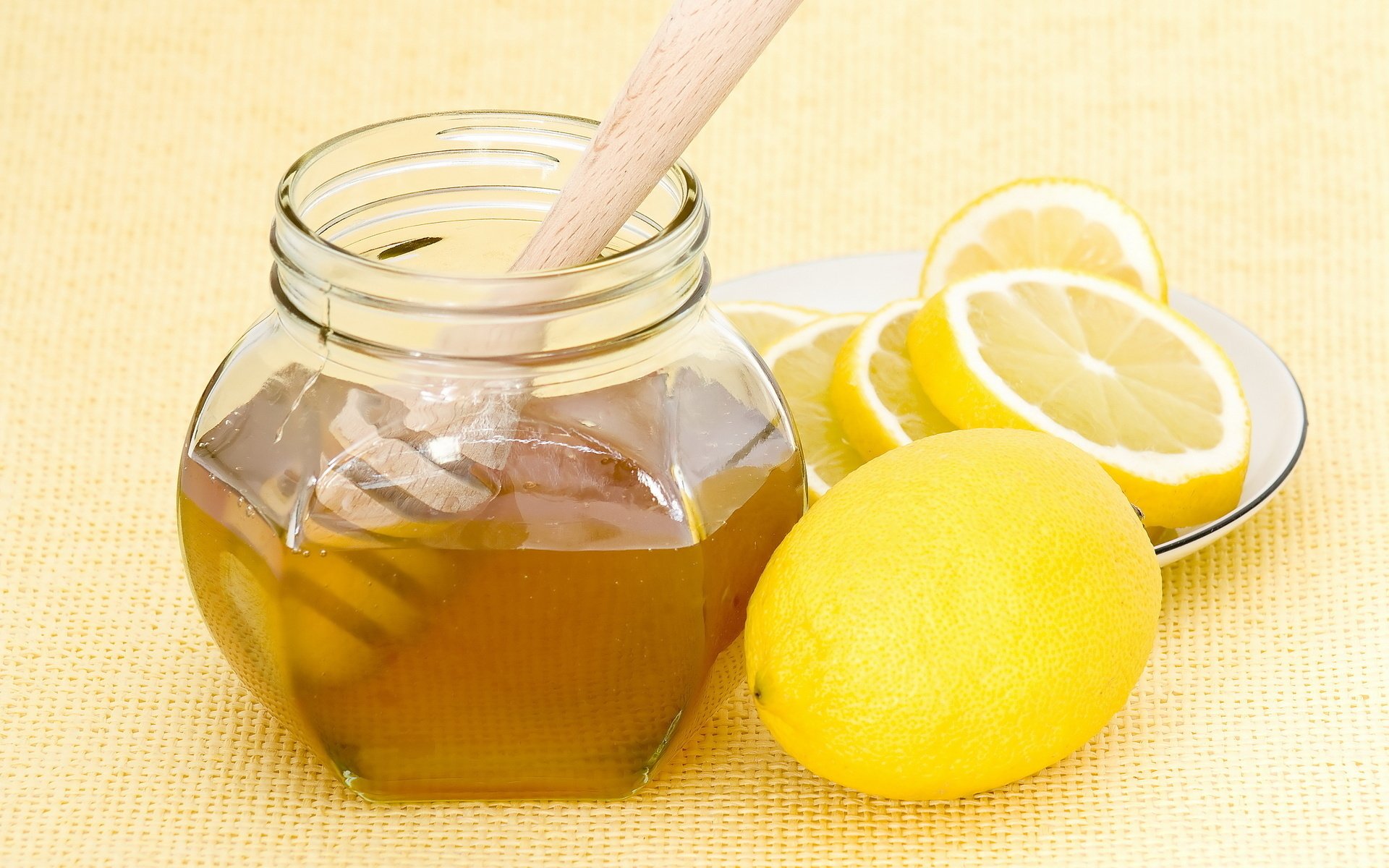 HD desktop wallpaper showing a jar of honey with a wooden dipper, accompanied by fresh whole and sliced lemons on a light yellow background.