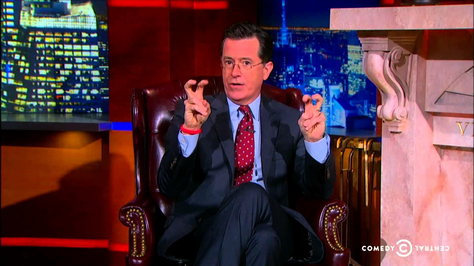 HD wallpaper of a man from The Colbert Report seated, making air quotes, with the show's set in the background.