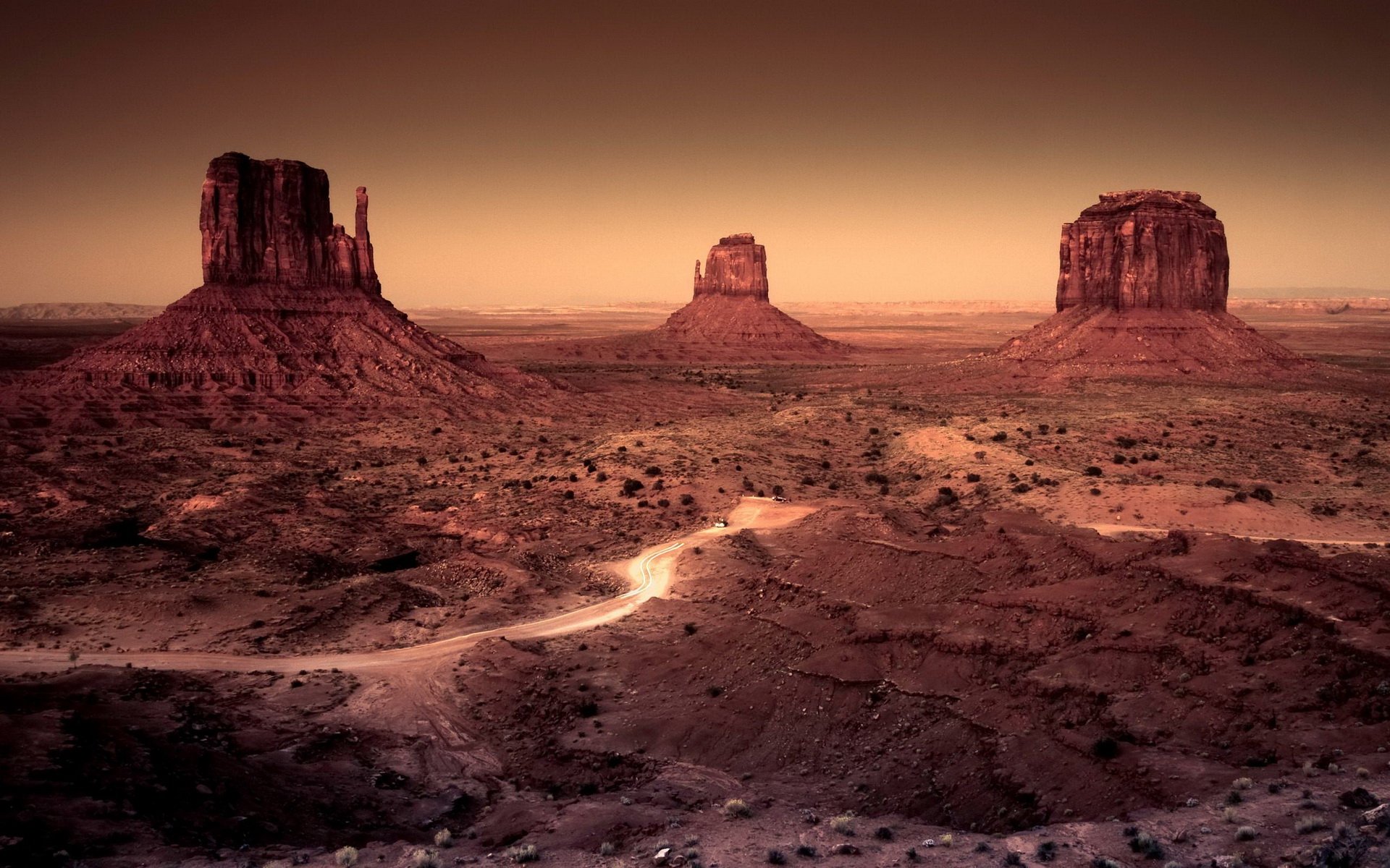 HD PC desktop wallpaper of a vast desert at sunset: three towering mesas over a cracked red-brown plain with a winding sandy trail leading into the horizon.