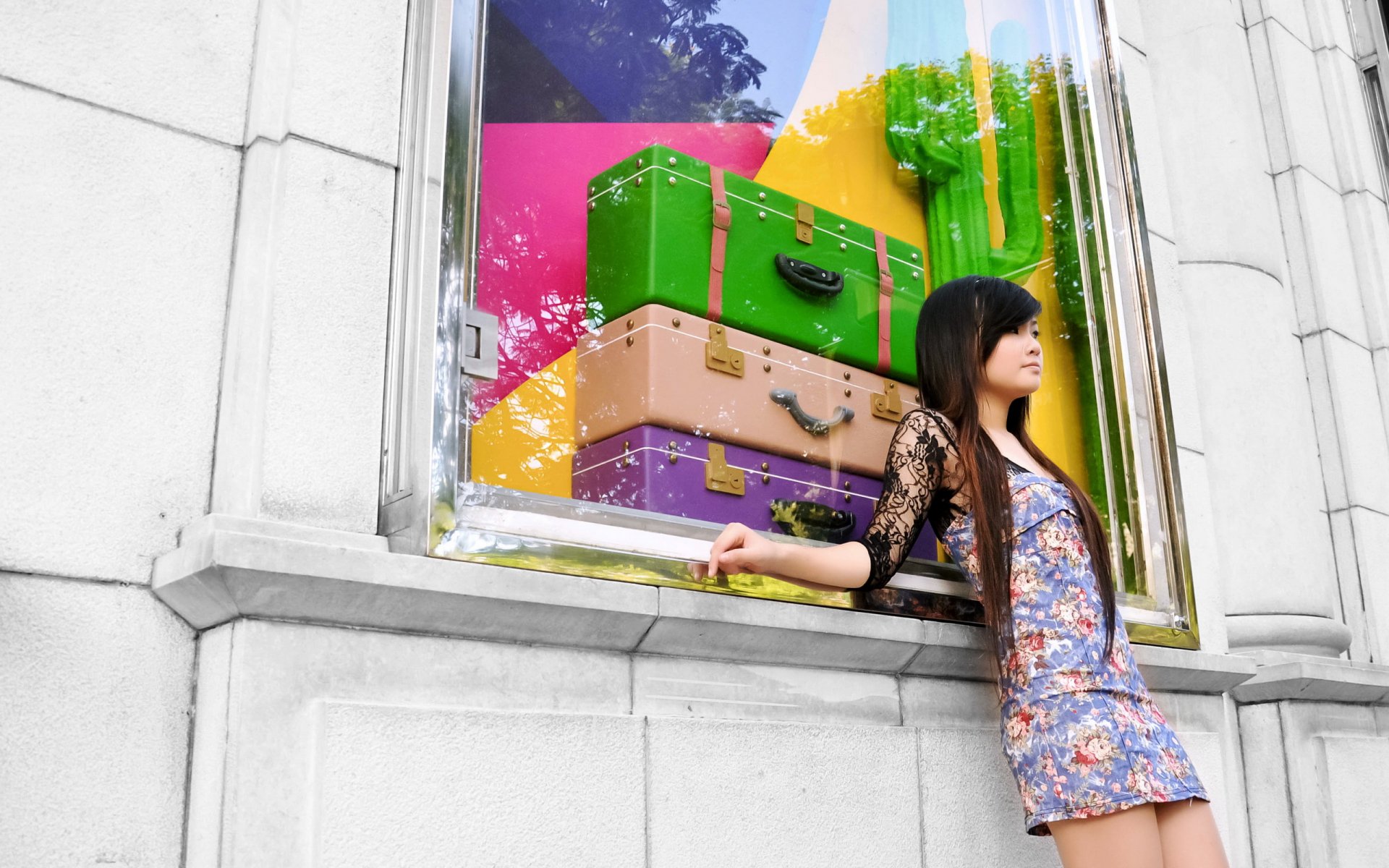 A woman with long hair leans against a colorful window display featuring stacked luggage and vibrant decor, creating an eye-catching HD desktop wallpaper.