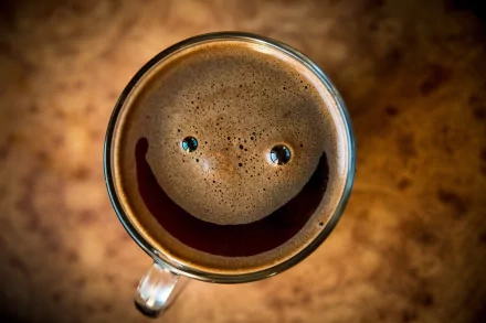 HD desktop wallpaper featuring a smiling cup of coffee with a foam smile and blue-eyed bubbles, set against a warmly toned background.