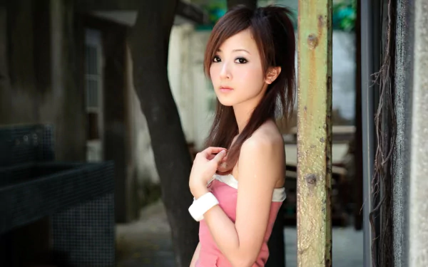 HD desktop wallpaper featuring a woman, Mikako Zhang Kaijie, standing outdoors against an industrial background. She gazes thoughtfully at the camera, adorned in a pink outfit with a white bracelet.