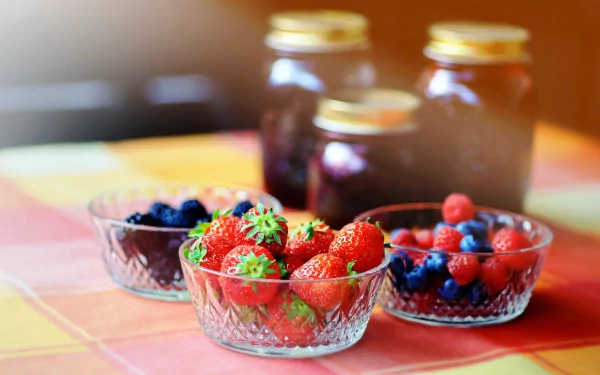 HD desktop wallpaper featuring vibrant bowls of fresh strawberries, blueberries, and raspberries with jars of preserves in the background on a checkered tablecloth.