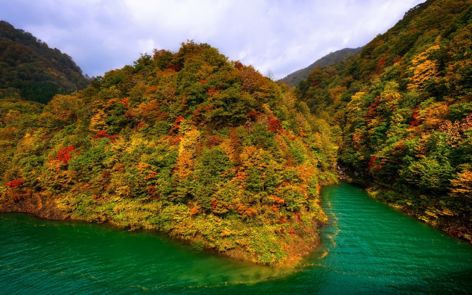 HD PC desktop wallpaper showing a vibrant fall landscape with dense orange and green trees surrounding a winding emerald river under a partly cloudy sky.