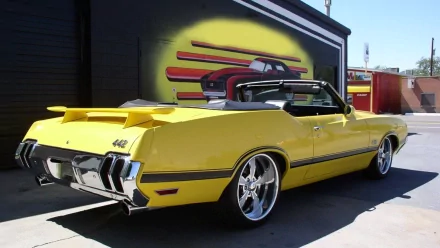 HD desktop wallpaper featuring a bright yellow Oldsmobile 442 convertible parked outdoors with a custom car mural in the background.
