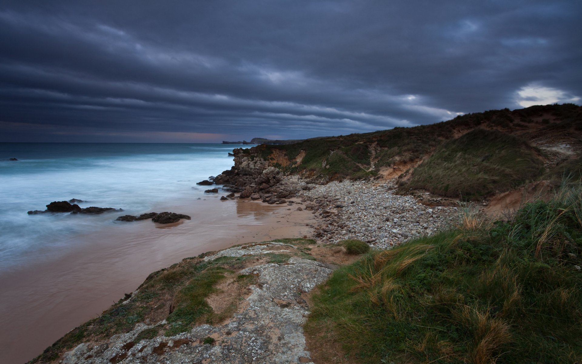 HD PC desktop wallpaper featuring a serene coastline with rocky shore, grassy cliffs, and moody clouds over a calm sea at dusk.