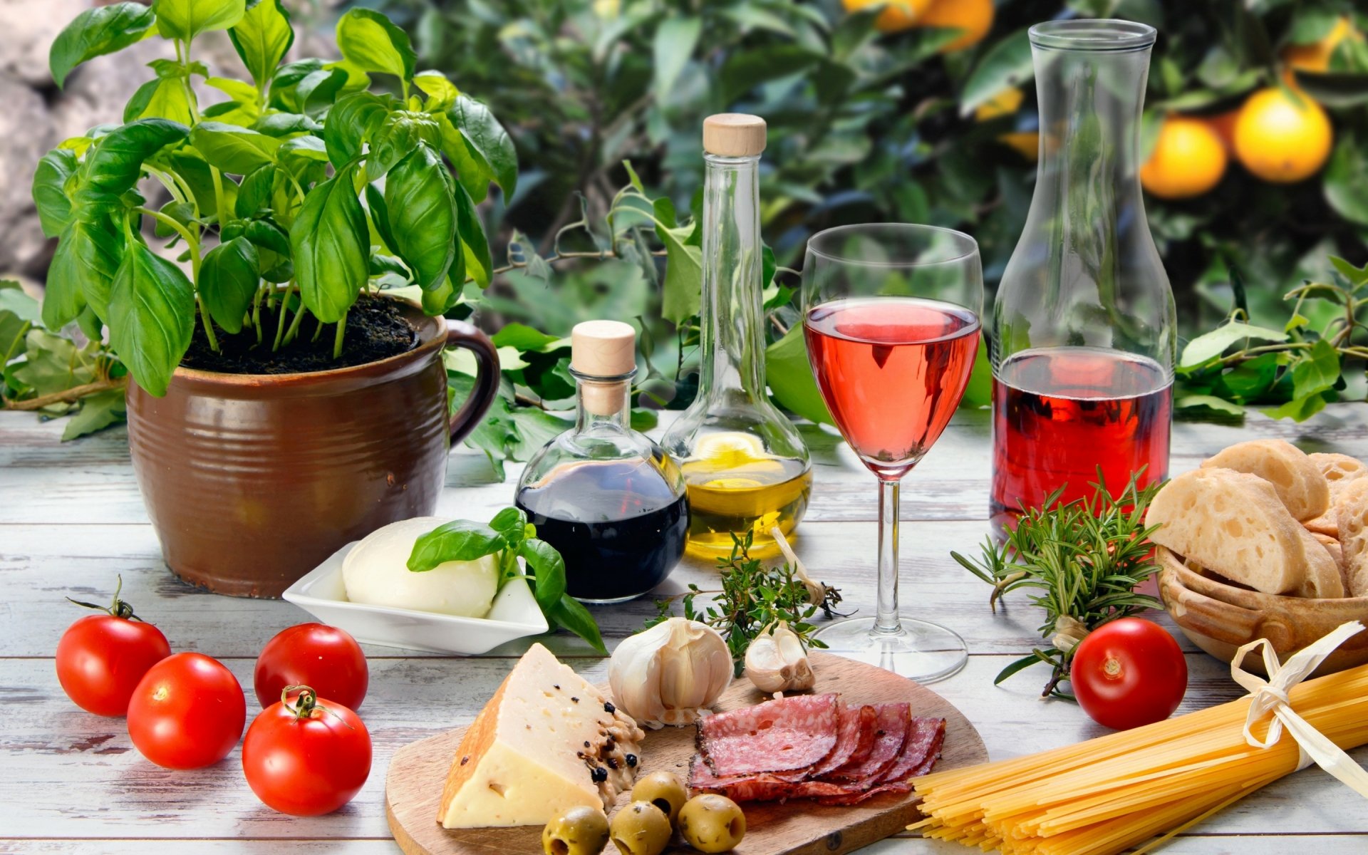 HD PC desktop wallpaper of a still life featuring fresh tomatoes, basil, garlic, cheese, cured meats, olives, pasta, and wine, set against a vibrant garden background.