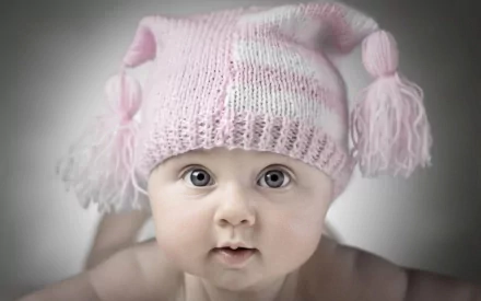A charming close-up of a baby wearing a pink knitted hat with tassels, capturing the innocence and wonder of early childhood. This high-definition image serves as a delightful desktop background.