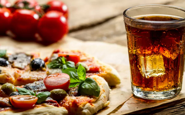 A delicious pizza topped with fresh vegetables sits on a wooden surface, accompanied by a glass of cola and ripe tomatoes, creating a vibrant HD desktop wallpaper.