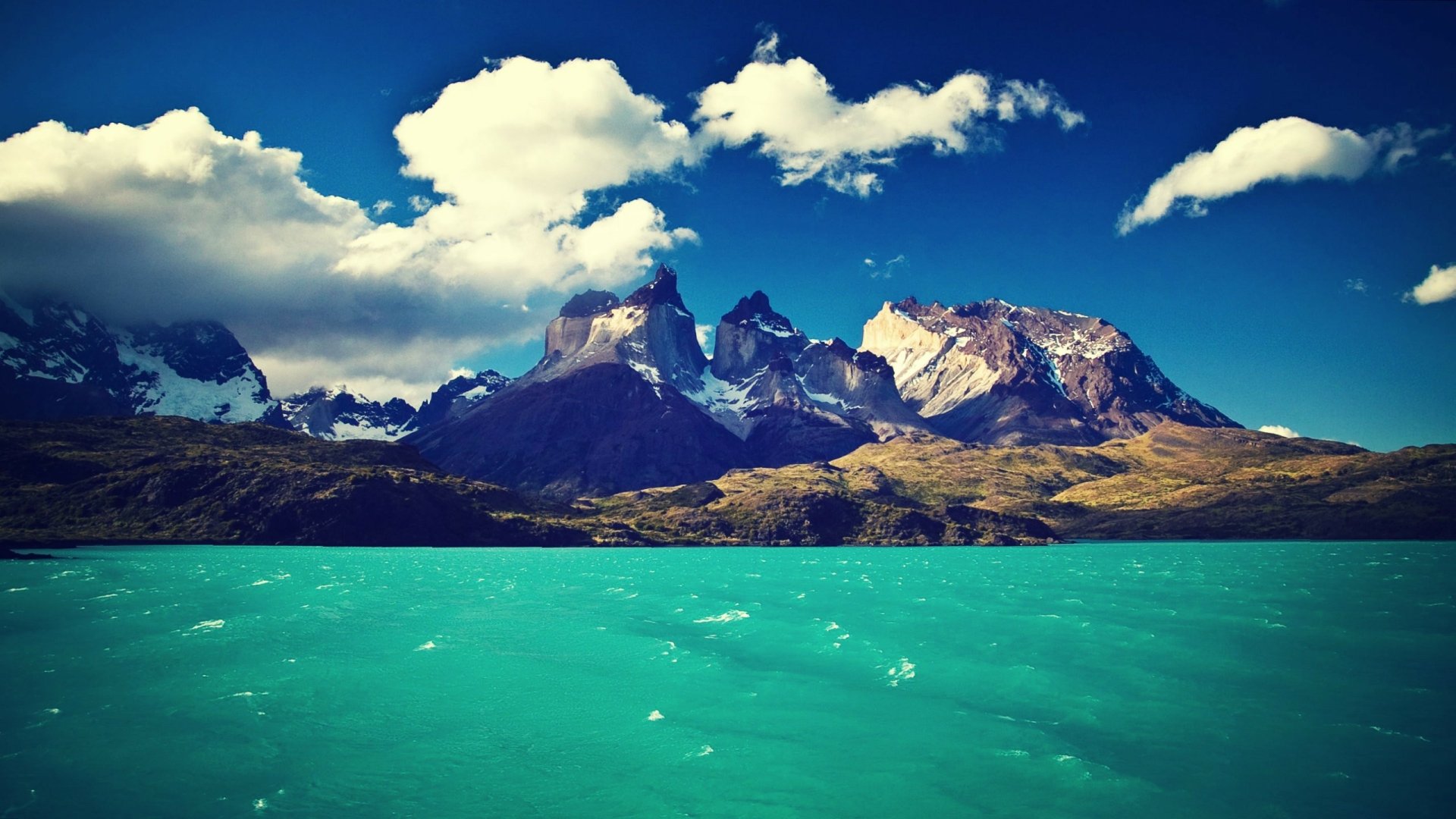 HD desktop wallpaper showcasing the striking turquoise waters and rugged peaks of Torres del Paine under a vibrant blue sky with scattered clouds.