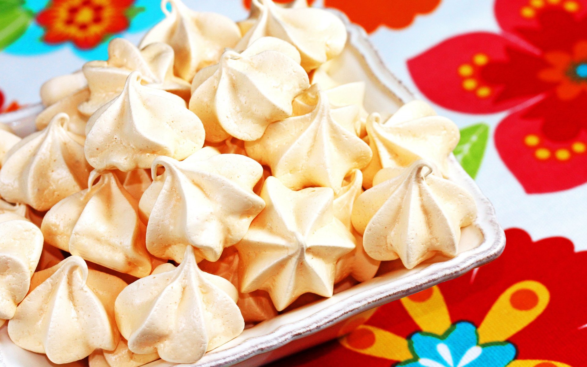 HD desktop wallpaper featuring a close-up of light, airy meringue cookies on a vibrant floral tablecloth background.