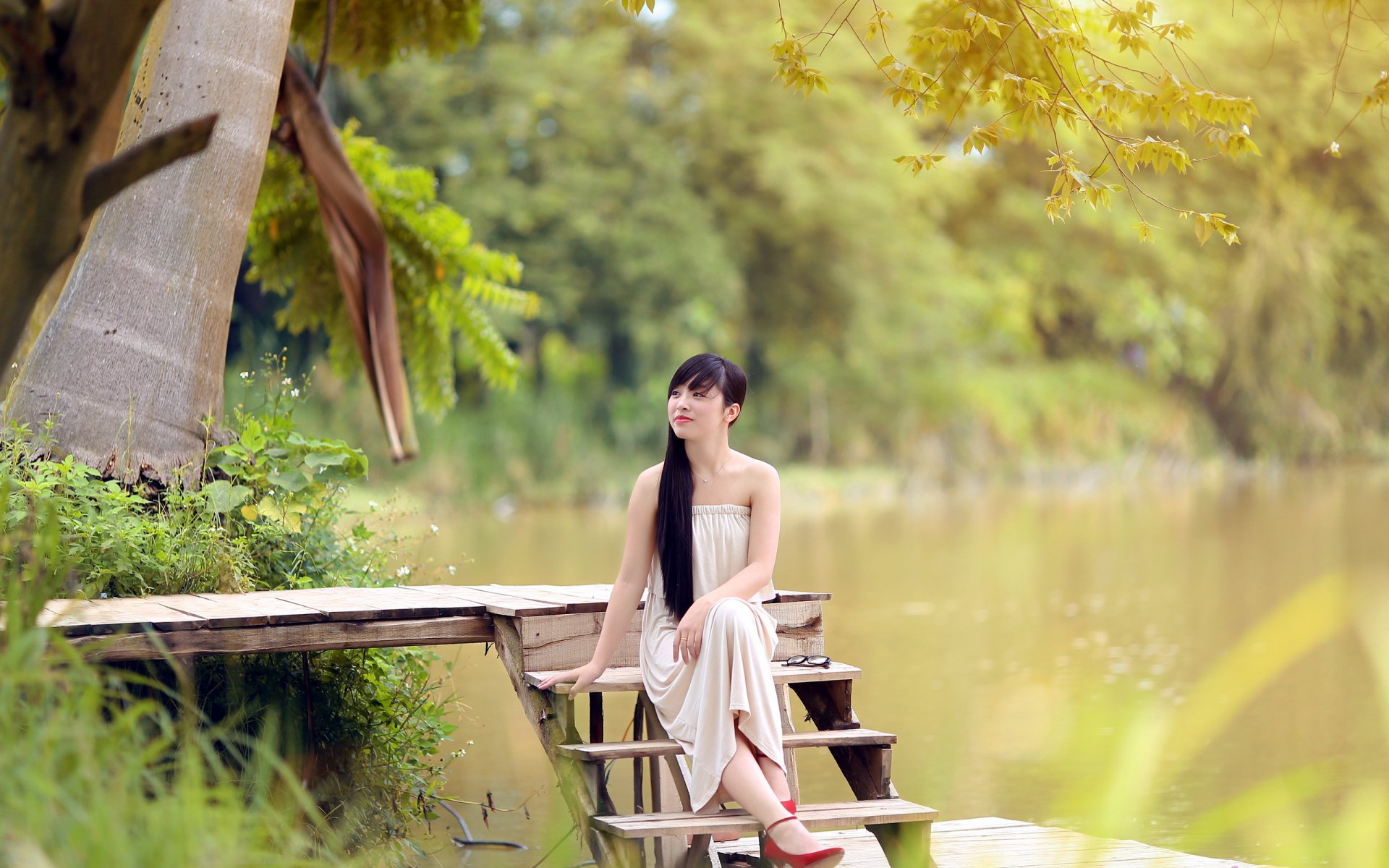HD desktop wallpaper featuring a serene Asian woman in a flowing dress sitting on a wooded dock beside a tranquil river, surrounded by lush greenery.