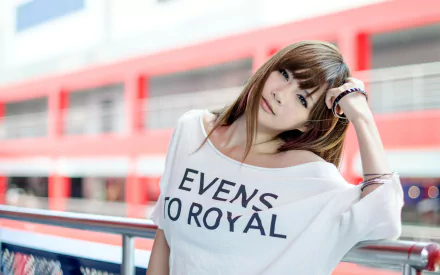 HD wallpaper featuring an Asian woman leaning on a railing, wearing a white shirt with EVENS TO ROYAL printed in black. The background shows a blurry red and white building.