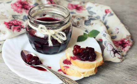 A jar of dark jam tied with twine sits beside slices of bread topped with jam, all arranged on a white plate with a floral napkin in the background. HD desktop wallpaper.
