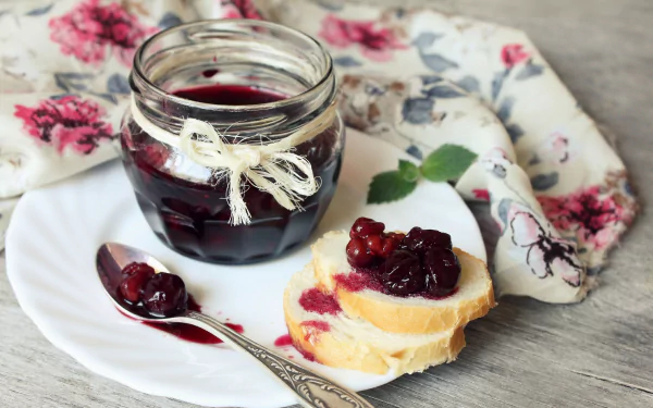 A jar of dark jam tied with twine sits beside slices of bread topped with jam, all arranged on a white plate with a floral napkin in the background. HD desktop wallpaper.
