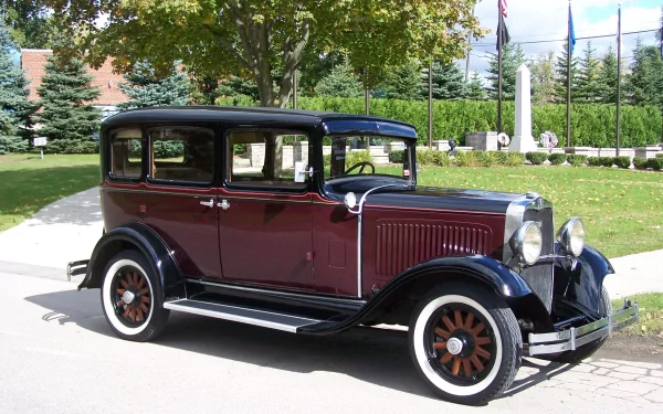 HD desktop wallpaper featuring a beautifully restored 1930 Dodge DC8 vehicle parked outdoors on a sunny day with trees and flags in the background.