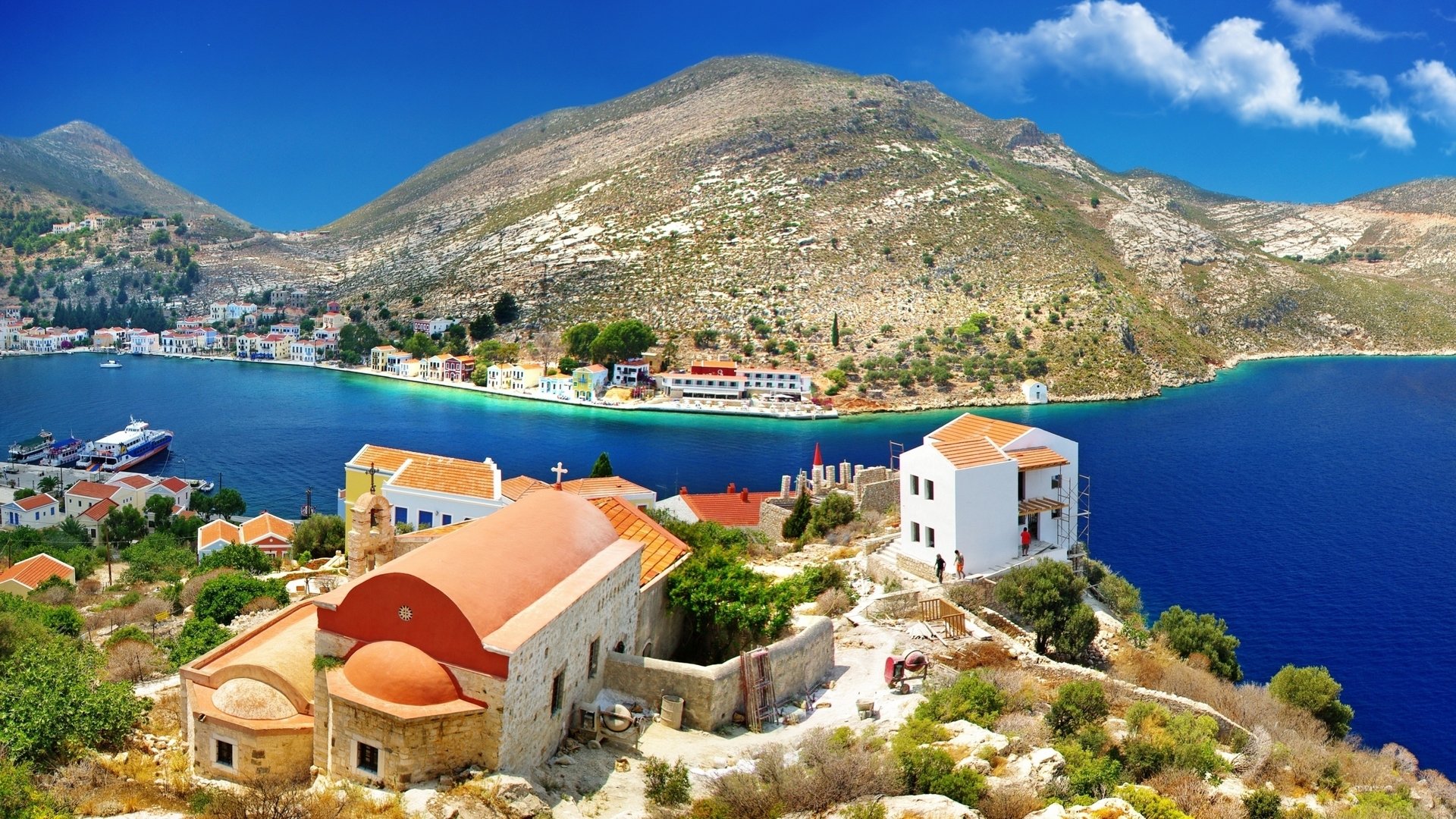 HD PC desktop wallpaper/background of Kastelorizo's man-made harbor and whitewashed buildings on a rocky shore, vivid blue sea and sunlit hillside.
