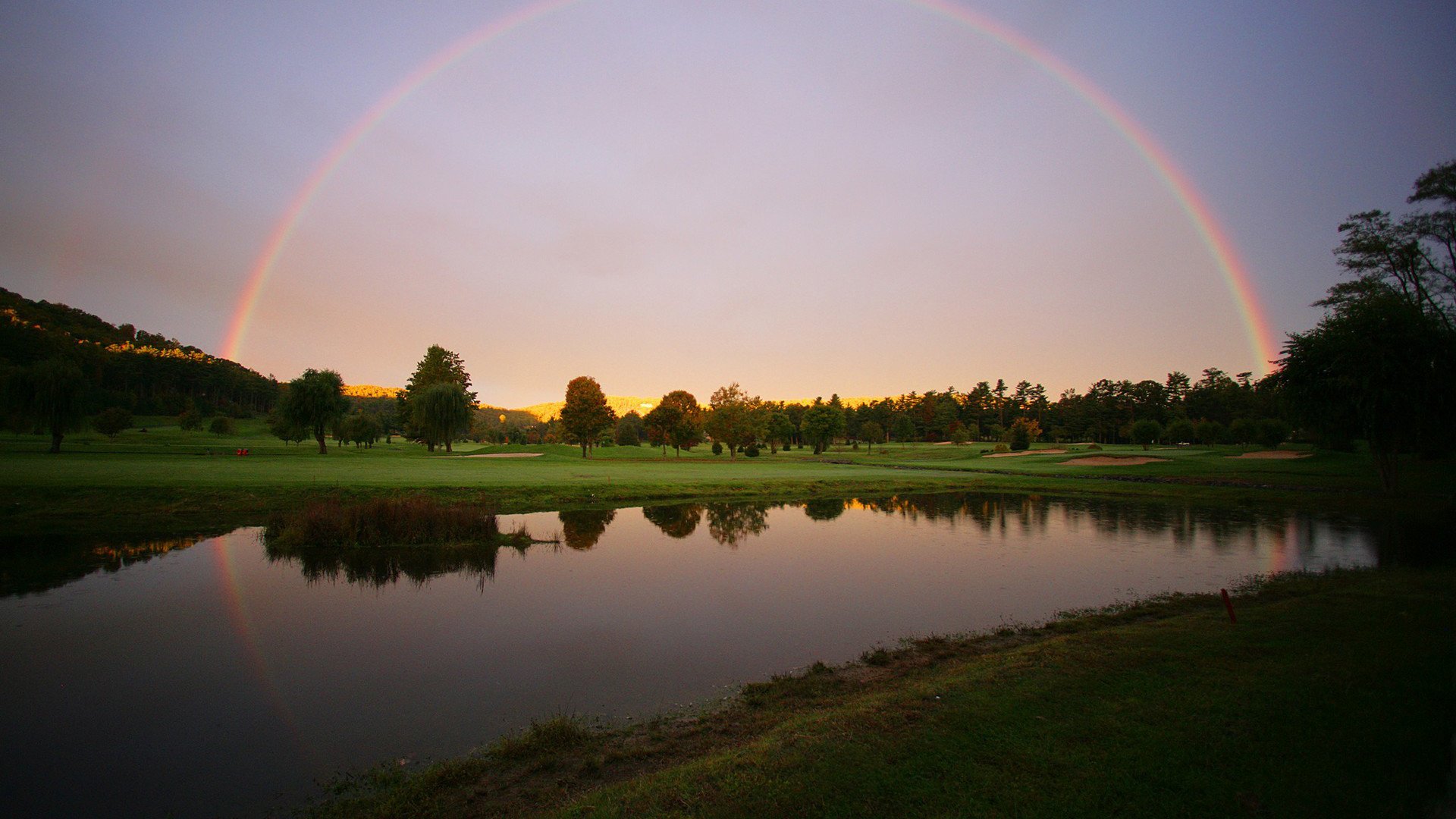 HD PC desktop wallpaper showing a serene lake under a glowing sky with a vibrant rainbow arching over a tree-lined horizon in nature.