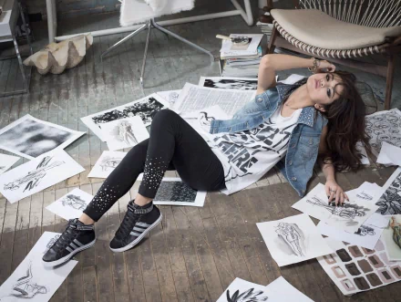 HD desktop wallpaper featuring Selena Gomez, a brunette with long hair and brown eyes, lounging on the floor surrounded by sketches and wearing a casual outfit. The theme blends her music and acting career.