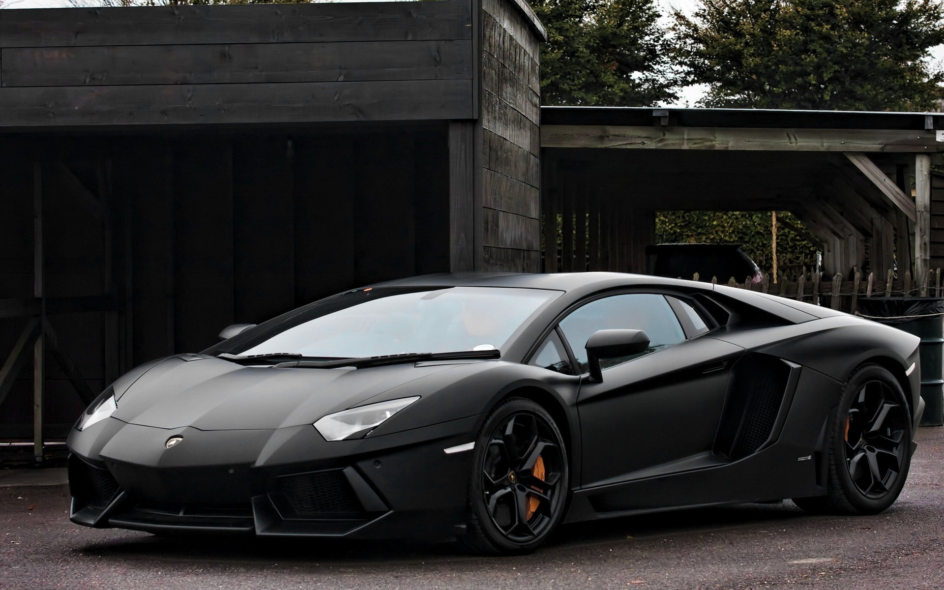 HD PC desktop wallpaper featuring a sleek black Lamborghini Aventador parked in front of a dark wooden structure with greenery in the background.