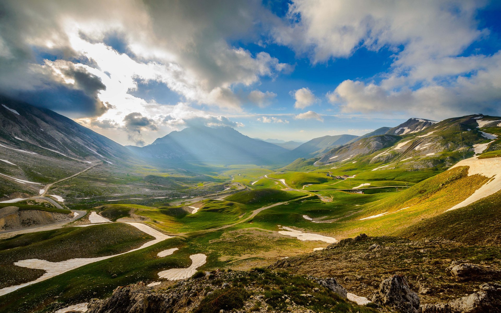 HD PC desktop wallpaper/background showing a nature landscape: sunlit alpine valley with winding roads, green meadows, snow patches and dramatic cloud rays over distant mountains.