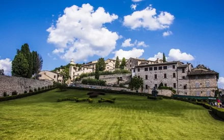 Man-made structures in Assisi under a bright blue sky with scattered clouds, featuring a lush green lawn in the foreground. HD PC desktop wallpaper and background.