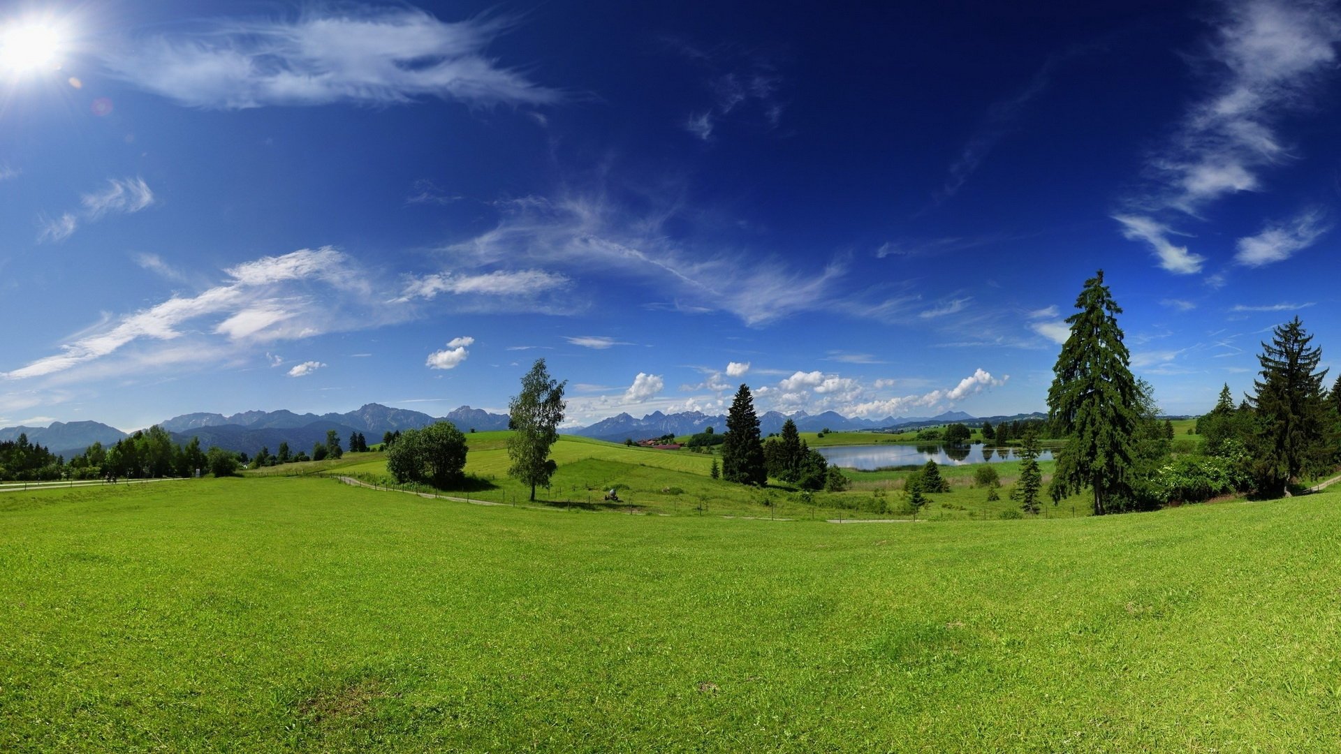 HD desktop wallpaper featuring a vibrant green landscape with trees, a lake, and distant mountains under a bright blue sky with scattered clouds.