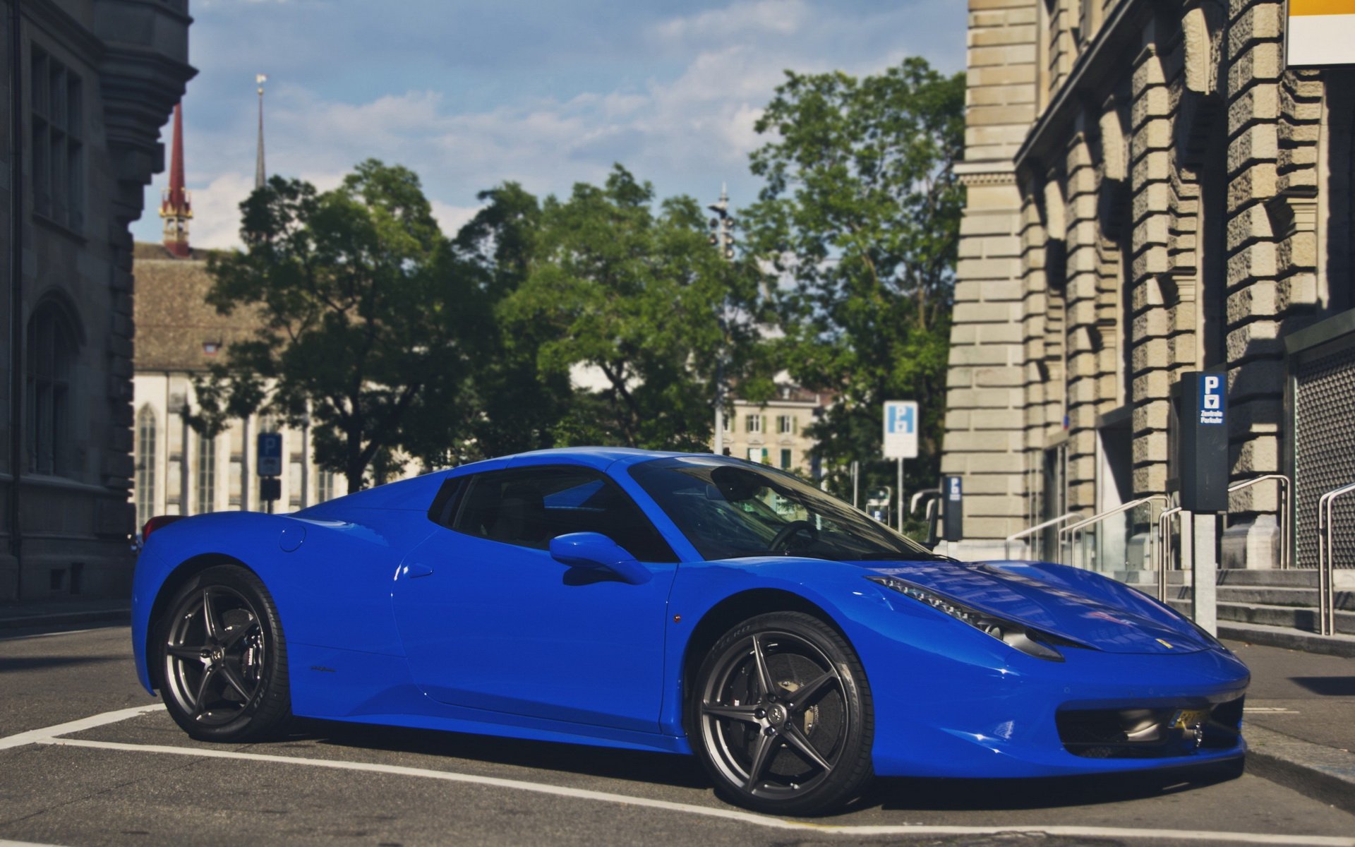 Blue Ferrari vehicle parked on a sunlit city street; HD PC desktop wallpaper/background showing classic stone buildings and leafy trees.