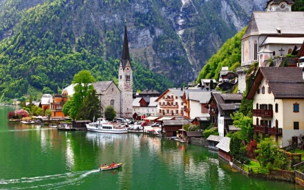 A scenic view of Hallstatt featuring a serene lake, charming houses, and a church with a tall spire, set against lush green mountains. This high-definition image serves as a beautiful desktop wallpaper.