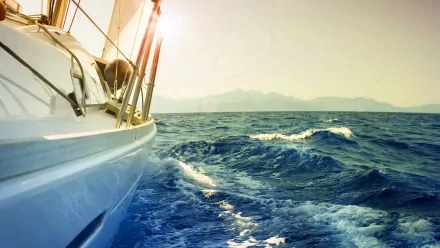 A sleek sailboat glides across sparkling blue waters, with soft waves and a warm sunset backdrop. This stunning image serves as a vibrant HD desktop wallpaper.