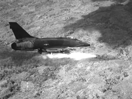 Black-and-white 2K Quad HD desktop wallpaper of a North American F-100 Super Sabre military jet flying low over rugged terrain, leaving a plume of rocket/exhaust smoke.