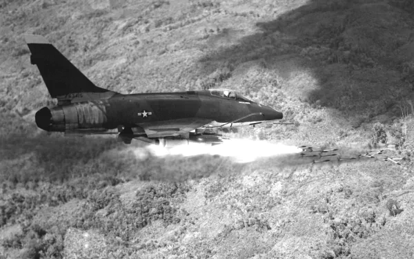 Black-and-white 2K Quad HD desktop wallpaper of a North American F-100 Super Sabre military jet flying low over rugged terrain, leaving a plume of rocket/exhaust smoke.