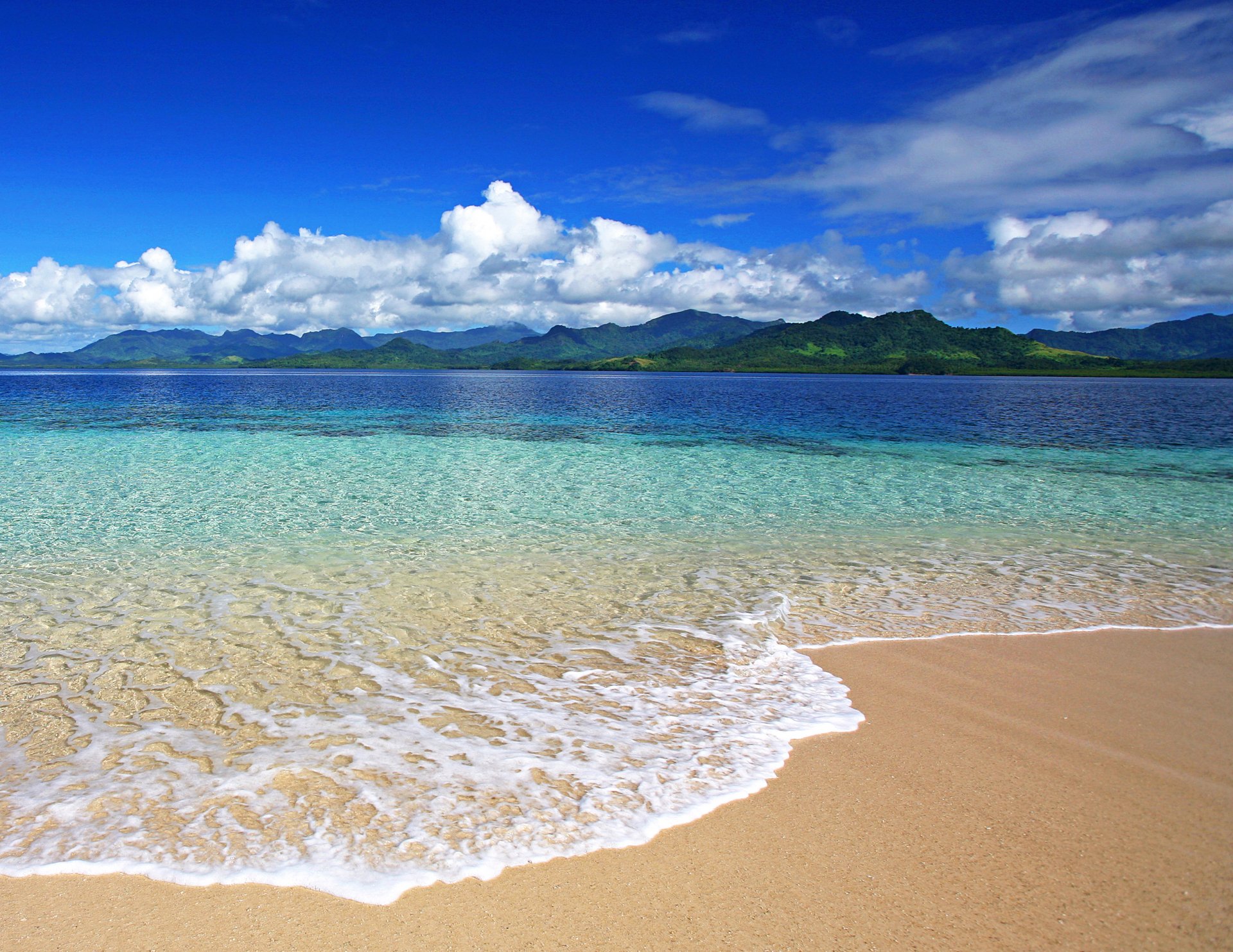 HD PC desktop wallpaper featuring a serene beach with clear turquoise water, gentle waves, and a distant mountain range under a bright blue sky with scattered clouds.