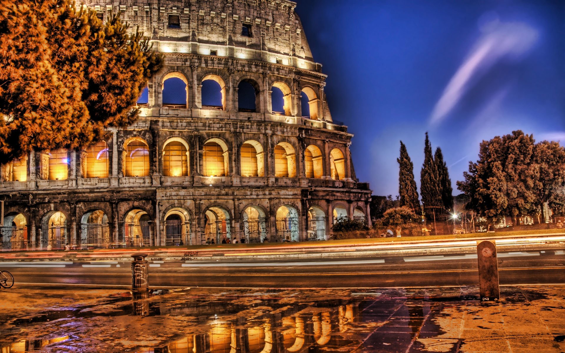 HD desktop wallpaper showcasing the illuminated man-made Colosseum at night with light trails and reflections on the wet pavement.