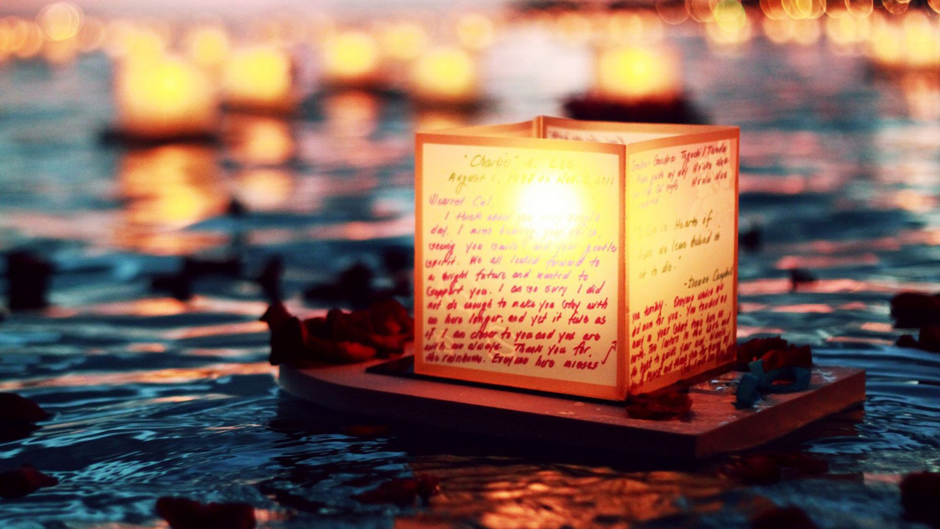 HD desktop wallpaper featuring a glowing man-made lantern floating on water with handwritten messages, surrounded by blurred lights creating a warm, serene atmosphere.