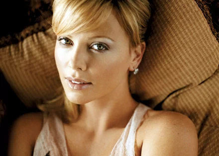 HD desktop wallpaper featuring celebrity Charlize Theron lying on a textured pillow, wearing a light-colored top and minimal jewelry.