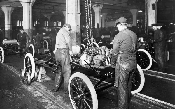 Black-and-white HD desktop wallpaper showing workers assembling a Ford Model T vehicle inside a factory.
