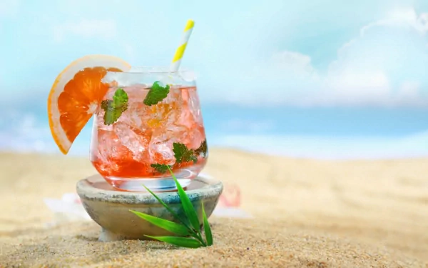 HD desktop wallpaper featuring a refreshing pink cocktail garnished with mint and a grapefruit slice, set on a sandy beach with a serene blue sky in the background.