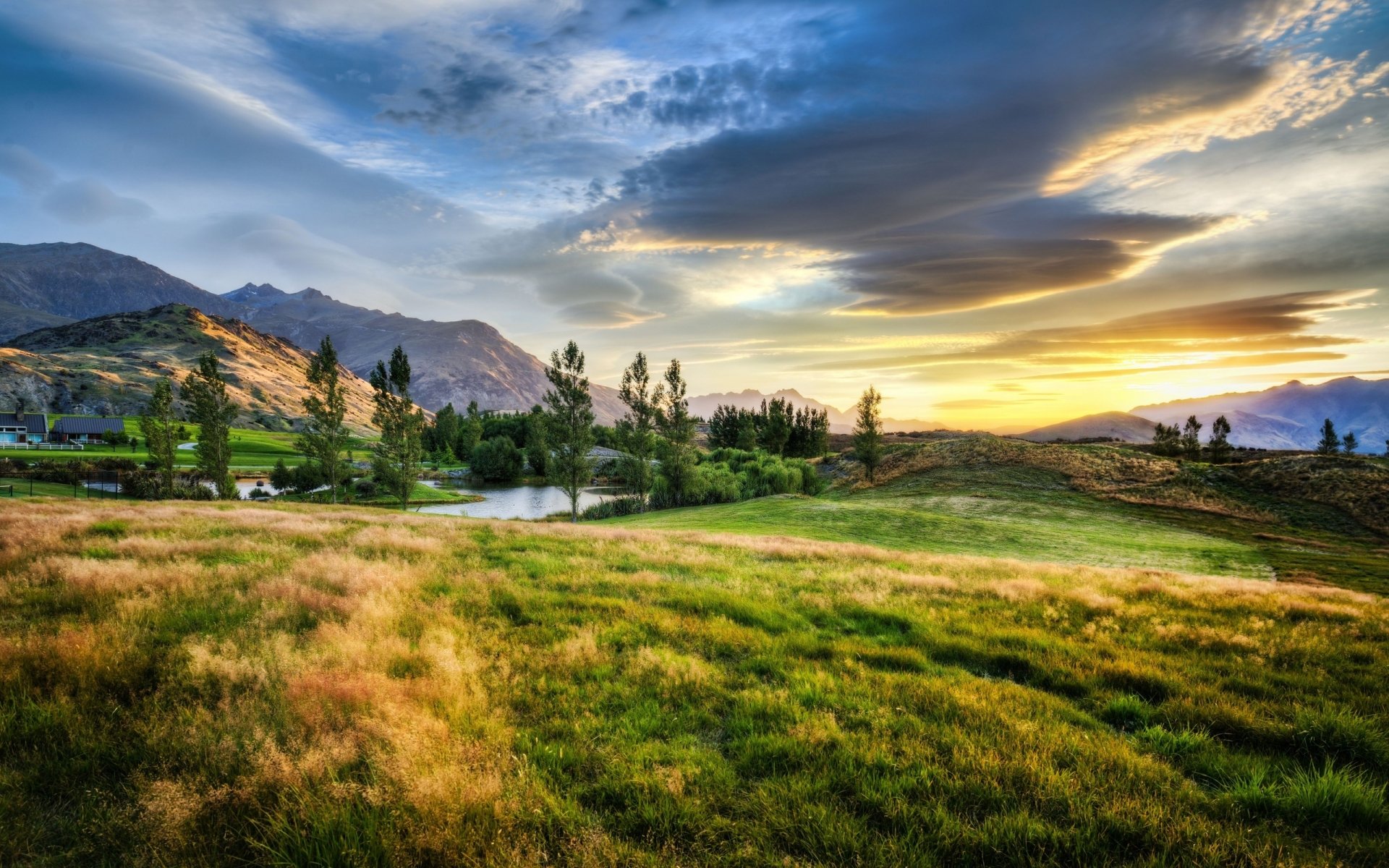 HD PC desktop wallpaper showcasing a vibrant nature landscape with rolling green hills, trees, distant mountains, and a dramatic sky at sunset.