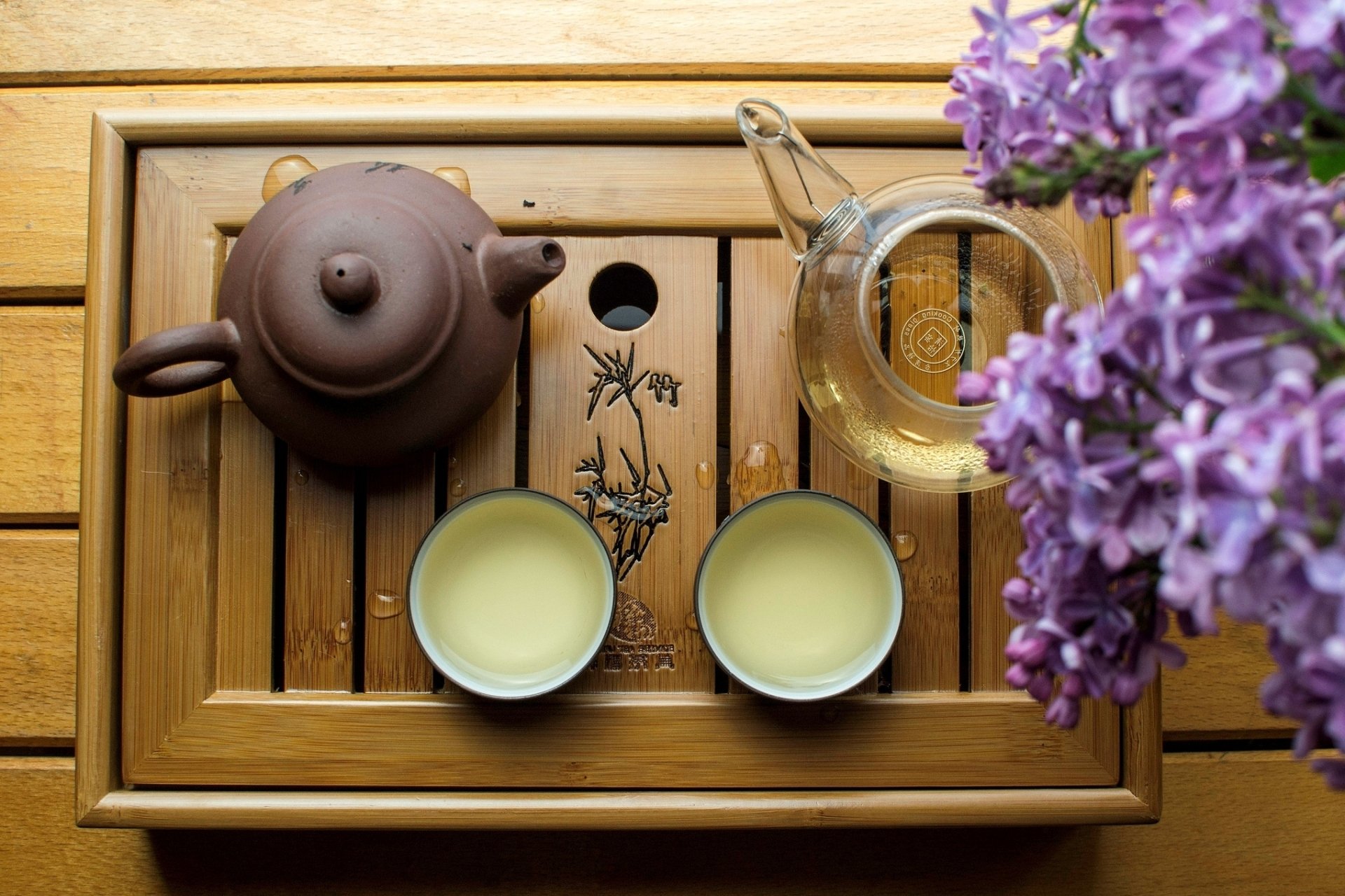 HD desktop wallpaper of a traditional tea set with a clay teapot, two cups of tea, a glass pitcher, and purple flowers arranged on a wooden tray.